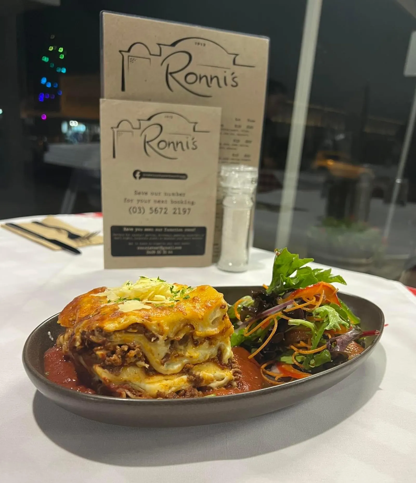 Classic lasagne bolognese with salad, two Ronni's menus propped behind on white tablecloth