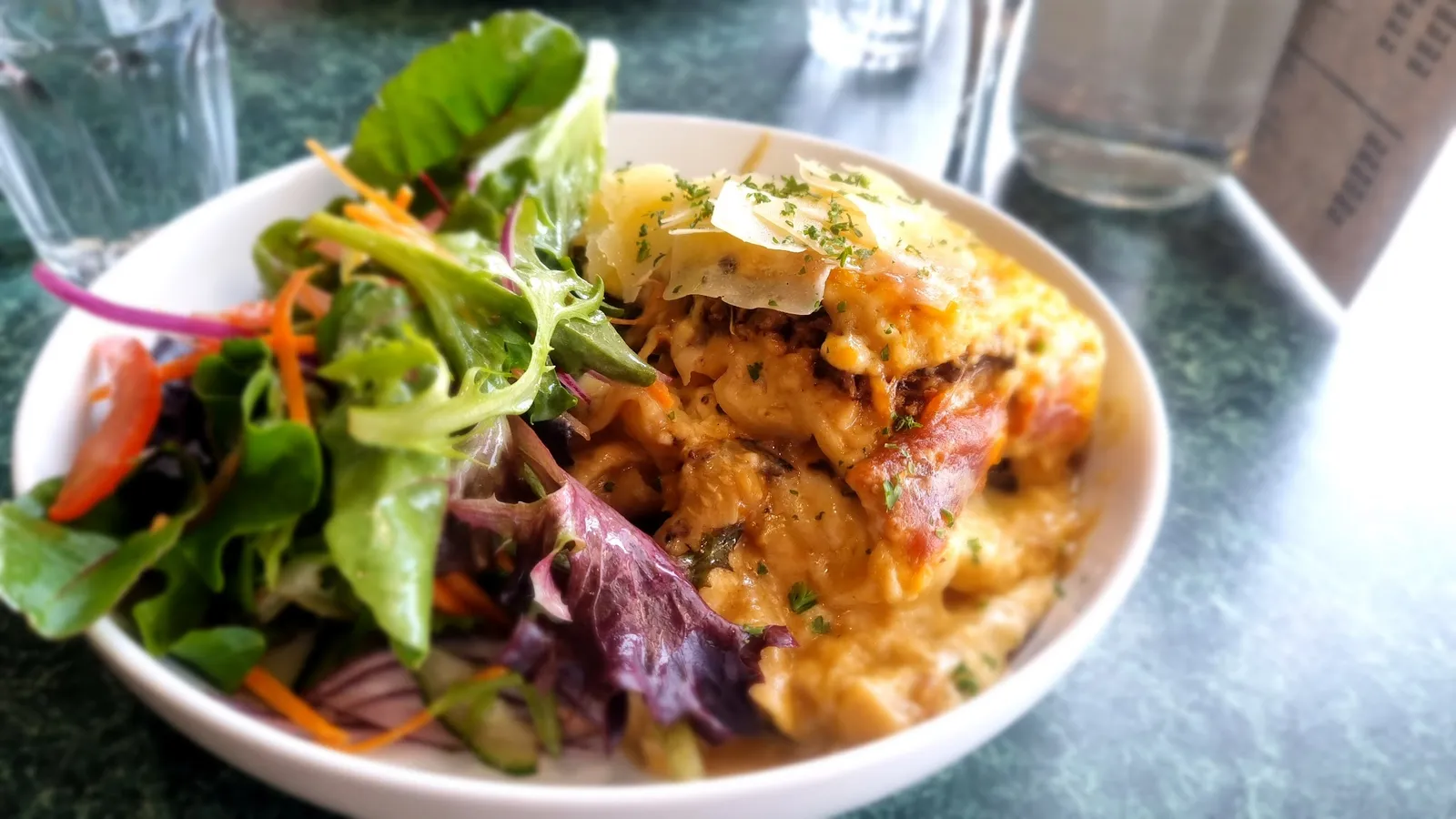 Lasagne with creamy cheese top and garden salad in white bowl on green marble